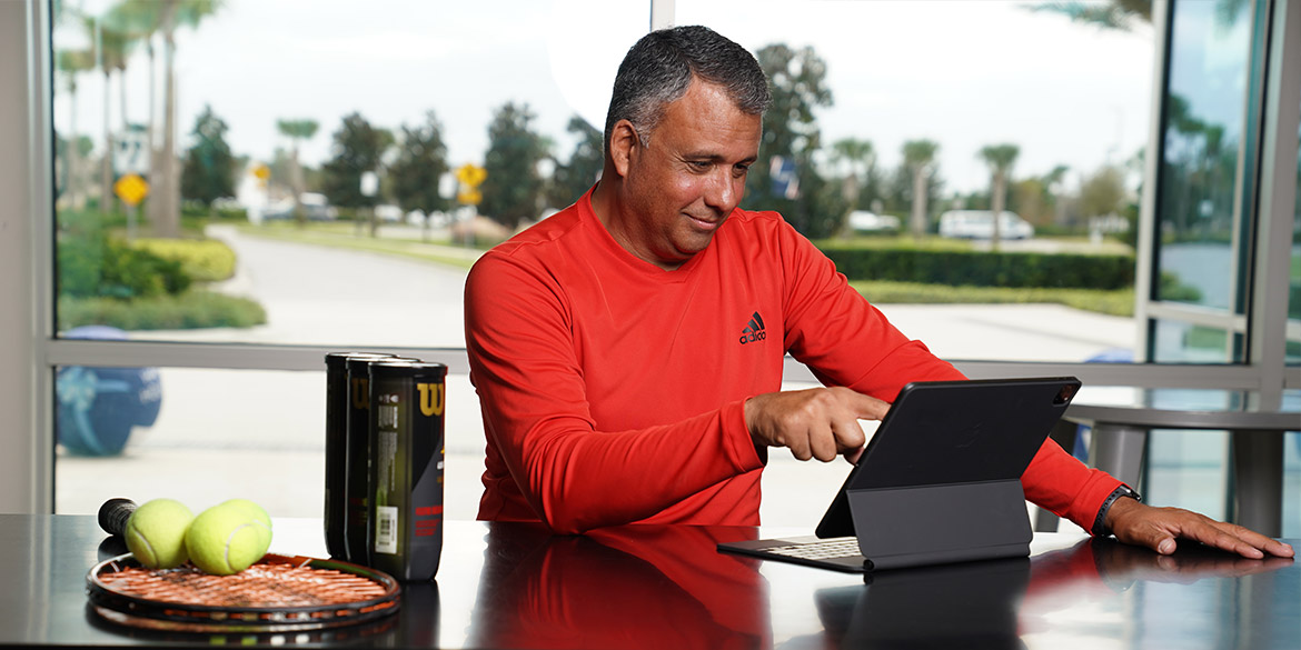 Older gentleman on laptop next to tennis balls and racquet.