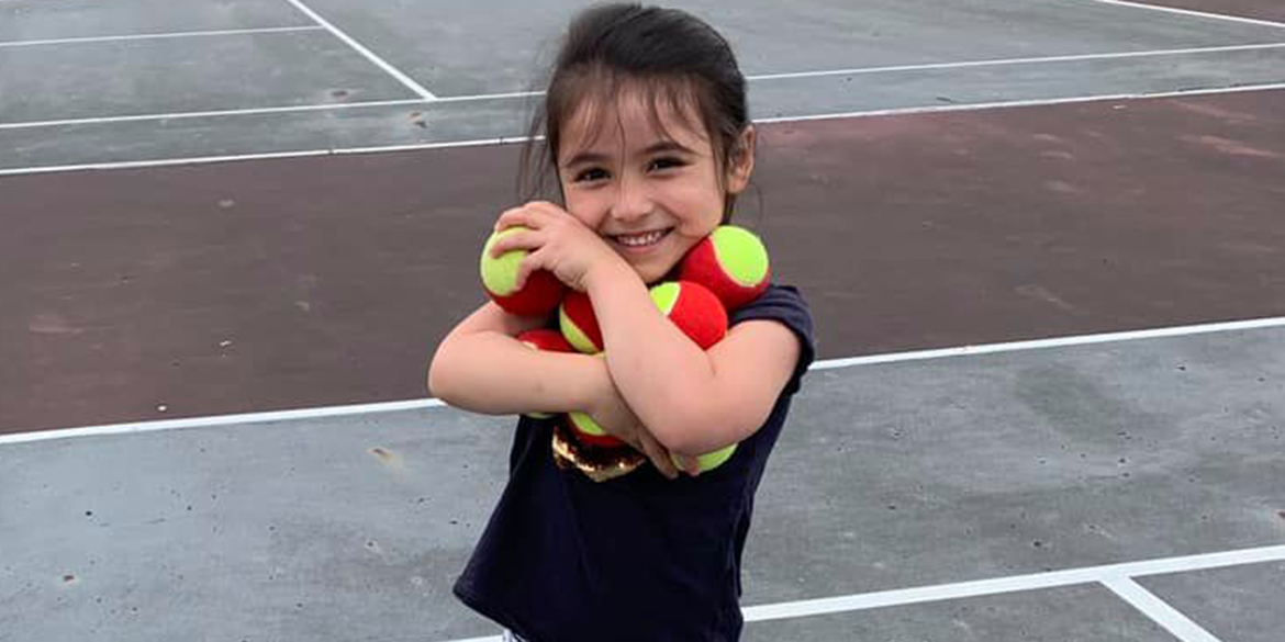 A young child stands on a tennis court holding red foam balls.