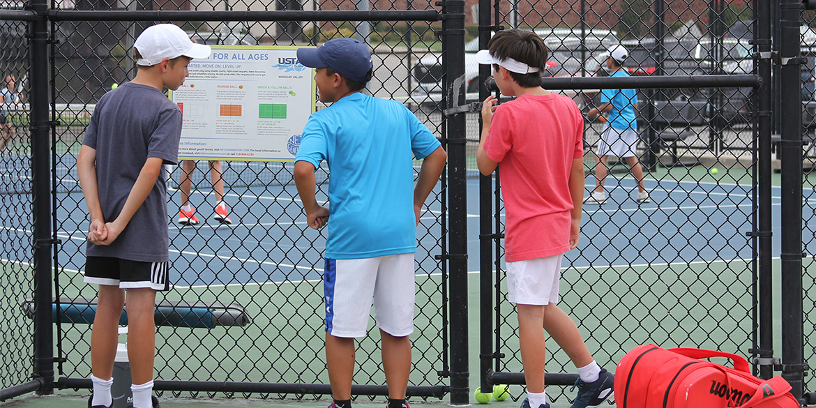 Three Junior Team Tennis players watch their teammates through a fence and look at a sign on the fence.